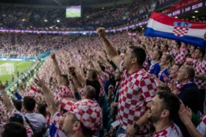 Croatian football fans singing and cheering in the stadium stands during a match, celebrating with flags and traditional Croatian football songs and chants